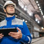 Trabalhador usando um tablet em um ambiente industrial, com equipamentos de segurança.