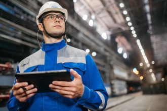 Trabalhador usando um tablet em um ambiente industrial, com equipamentos de segurança.