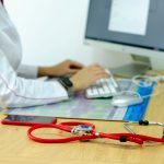 Profissional de saúde digita em um computador ao lado de um estetoscópio, em um ambiente clínico.