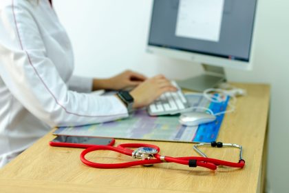 Profissional de saúde digita em um computador ao lado de um estetoscópio, em um ambiente clínico.
