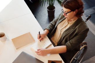 Profissional em cadeira de rodas escreve anotações em uma mesa de escritório moderno.