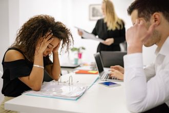 Mulher demonstra estresse diante do computador em um escritório, enquanto colegas trabalham ao fundo.