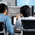 Dois desenvolvedores colaboram em um escritório enquanto analisam código exibido em uma tela.