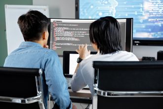 Dois desenvolvedores colaboram em um escritório enquanto analisam código exibido em uma tela.